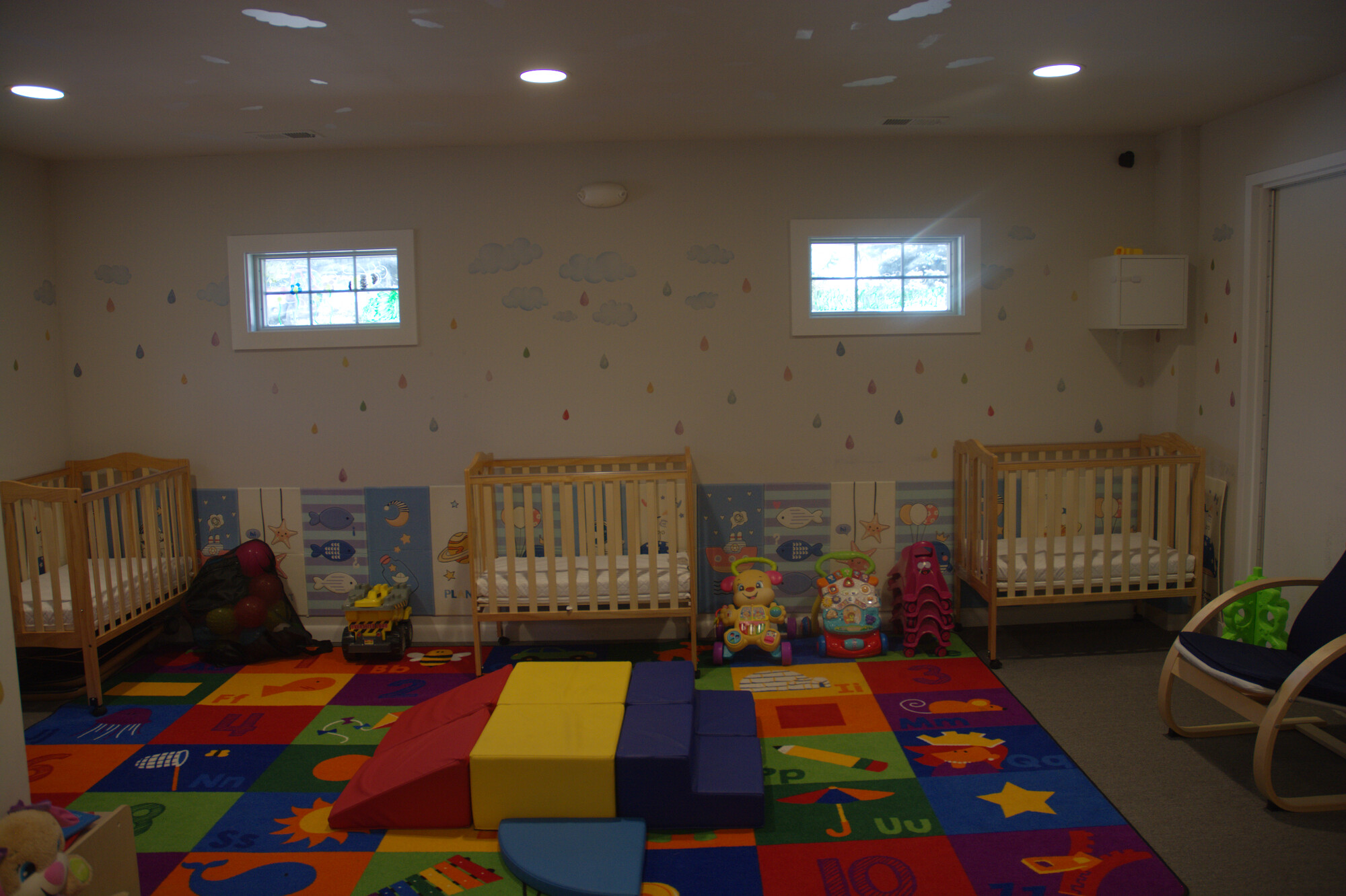 Infant room with cribs, cloud mural, and a colorful alphabet play mat